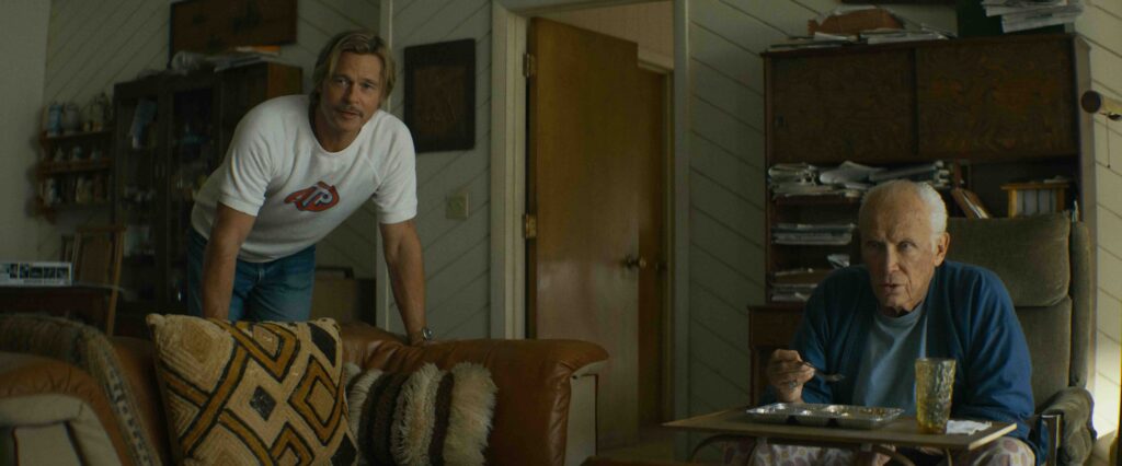 "Two men in a cozy living room, one sitting and eating at a tray table, the other leaning on a couch. The setting is warm with patterned pillows, wooden furniture, and shelves."